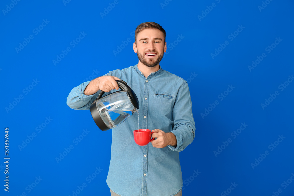 Young man pouring hot boiled water from electric kettle into cup on color background