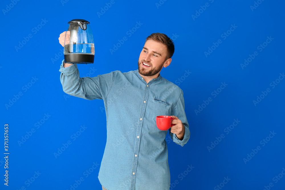 Young man with electric kettle and cup on color background