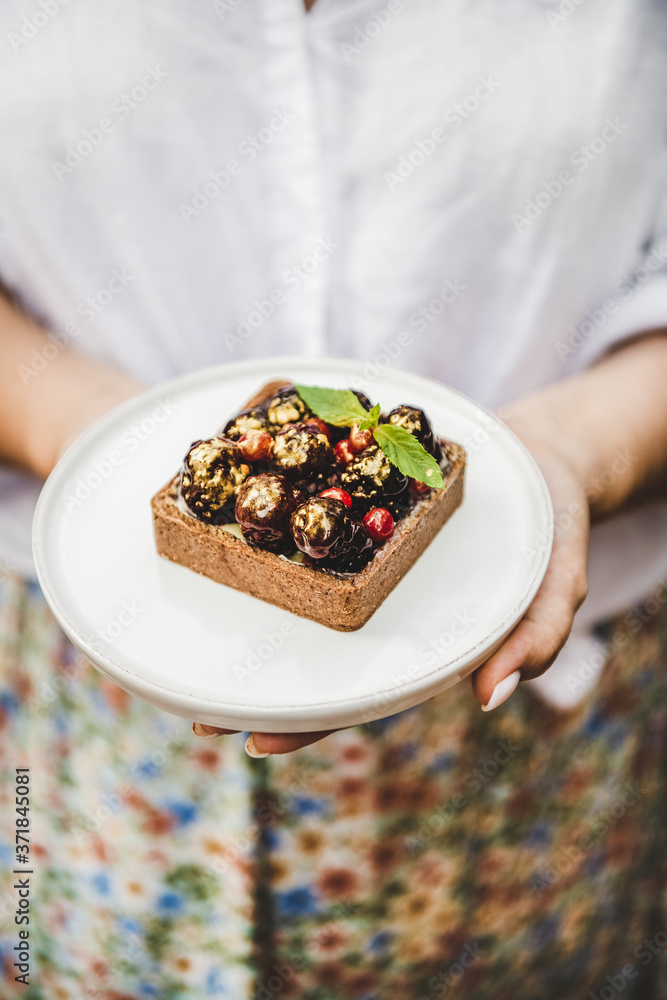 Woman holding cocoa tartalette with fresh seasonal berries on white plate in hands, selective focus.