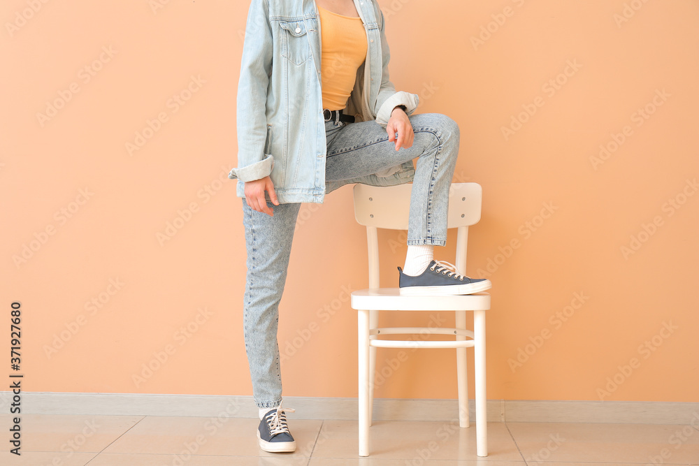 Beautiful young woman in jeans clothes and chair near color wall