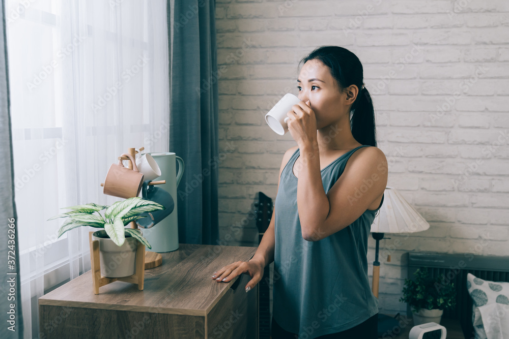 亚洲运动型女人站在窗户旁，把手放在橱柜上，锻炼后正在喝酒。下巴