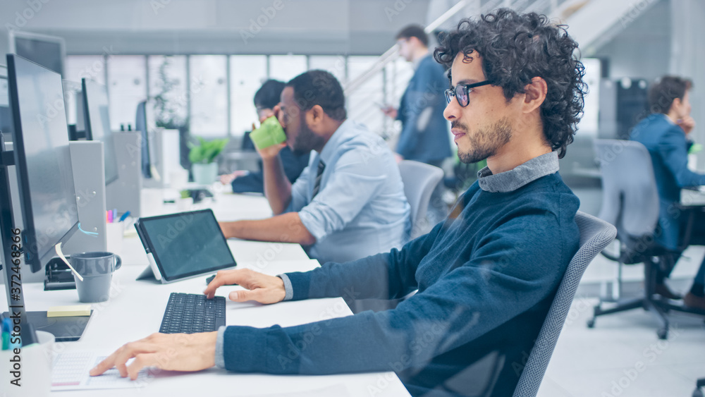 Young Handsome Manager with Curly Hair Works on a Desktop Computer and Compares Data with a Software