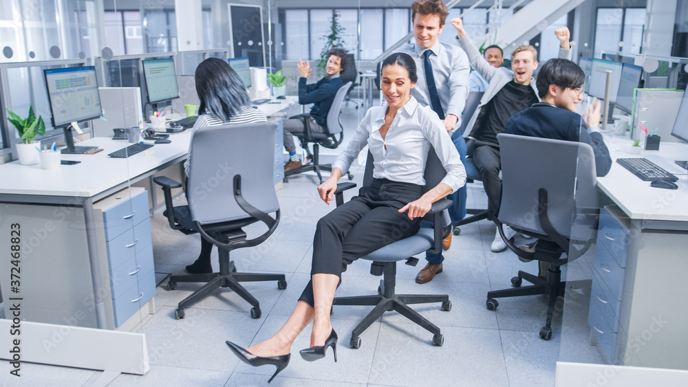 Cheerful Employee Pushes His Beautiful Female Colleague on a Chair Between Rows of Desks with Divers