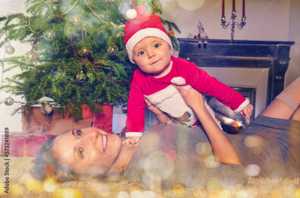 Mother lay on the ground with little baby toddler boy in front of the Christmas tree smiling and loo