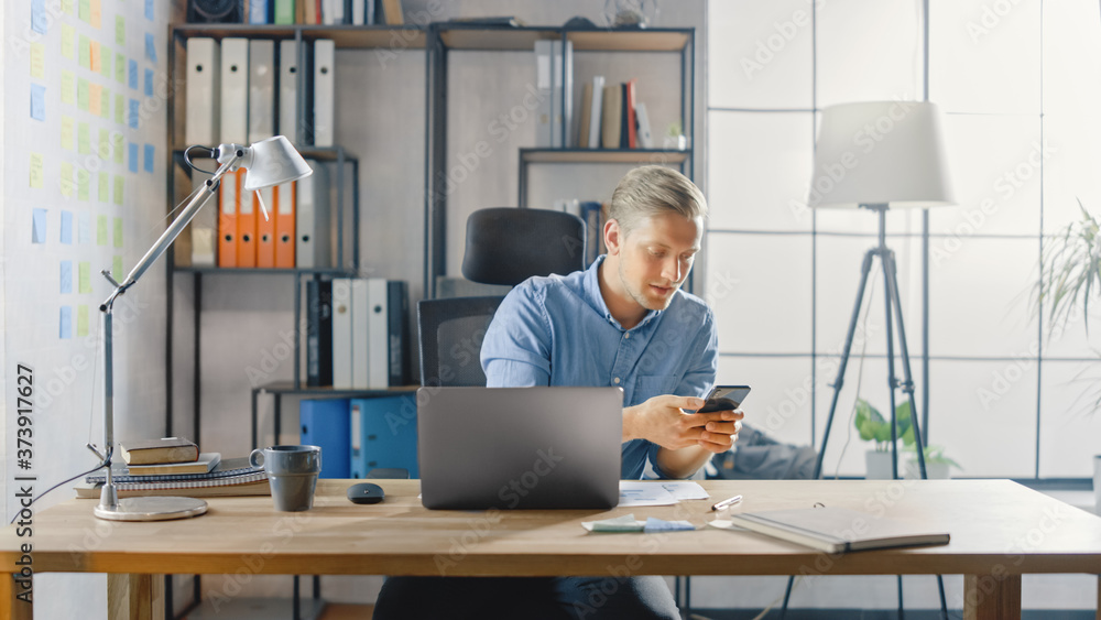 Entrepreneur Sitting at His Desk Works on Desktop Computer in the Stylish Office, Checks His Smartph