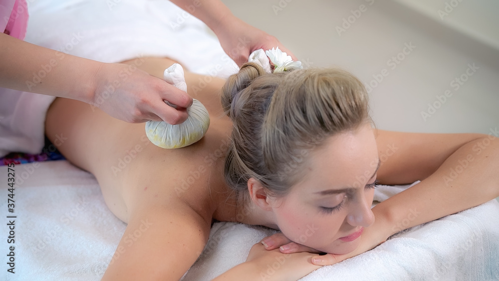 Massage therapist holds a herbal compress to do treatment to woman lying on spa bed in a luxury spa 