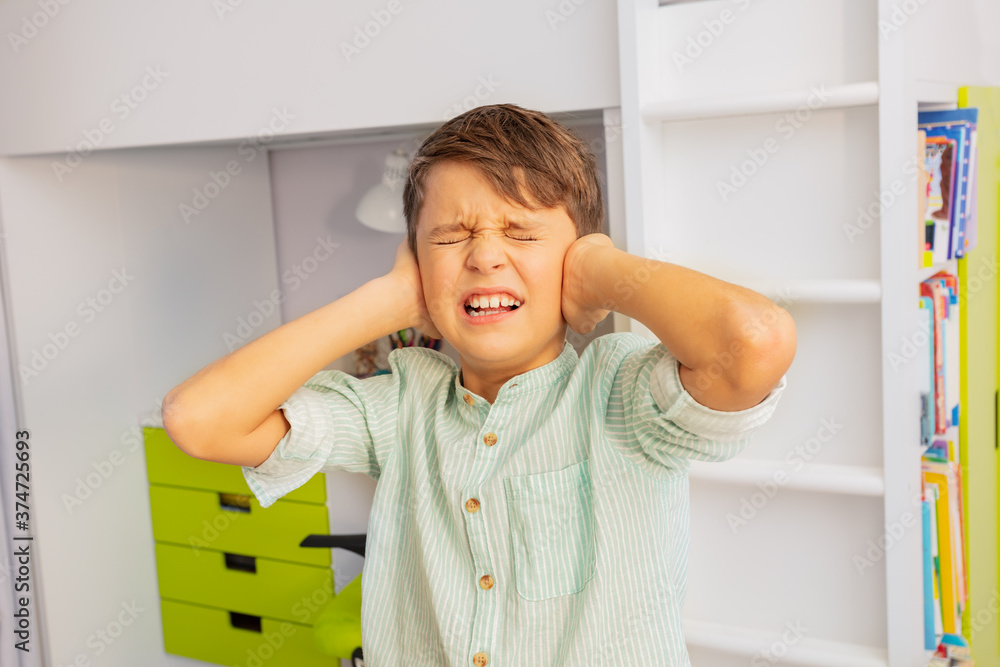 Close portrait of little autistic boy with strong negative face expression closing ears and grins te