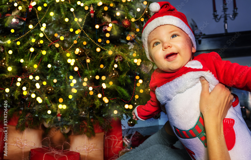 Happy little toddler baby boy in front of Christmas tree in mothers hands