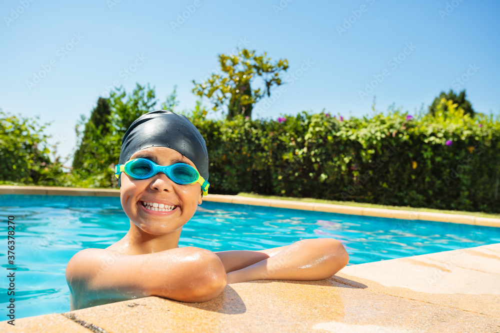 Happy little boy in swimming cap and googles on the border of outdoor pool look at camera with smile