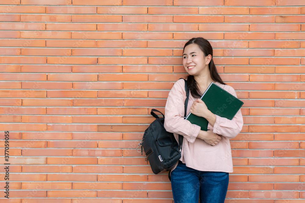 Asian smiling woman student holding book posing on brick background in campus. Happy teen girl high 