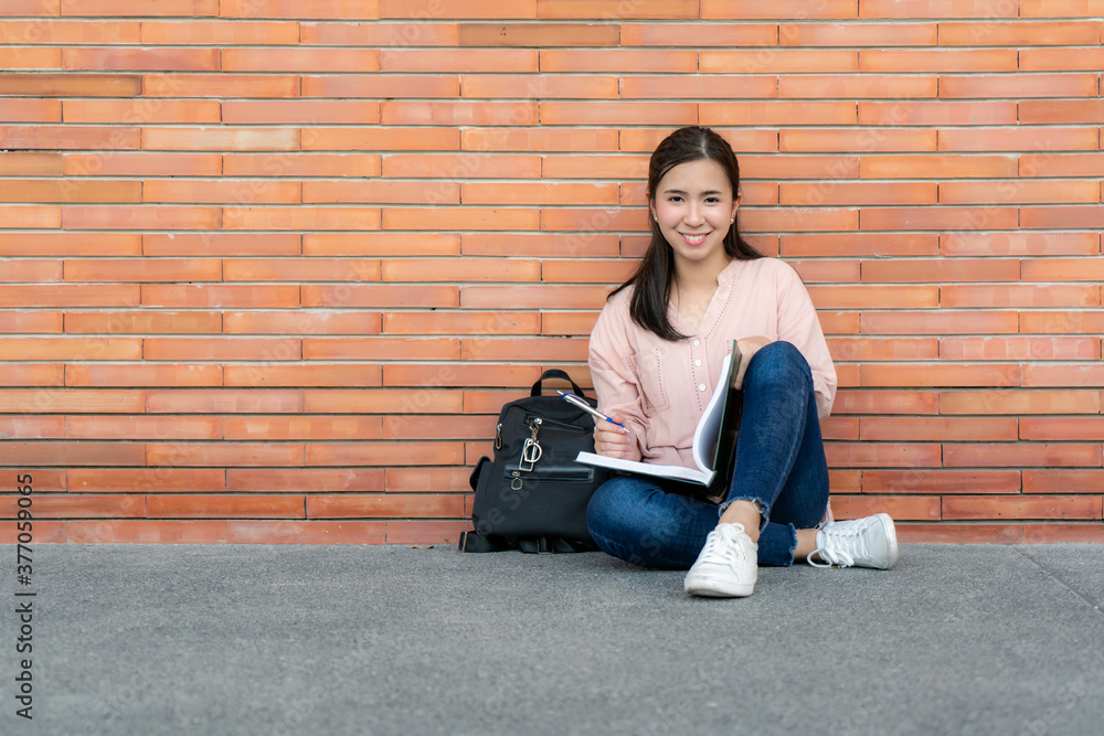 Asian smiling woman student holding book posing on brick background in campus. Happy teen girl high 