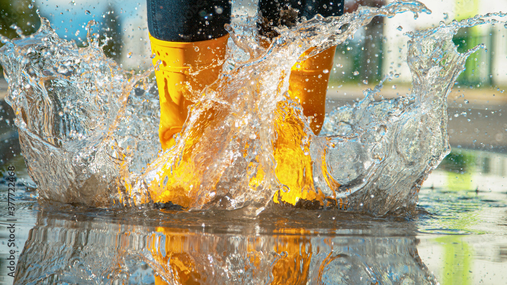 CLOSE UP Unrecognizable girl in yellow rubber boots jumps into the glassy puddle