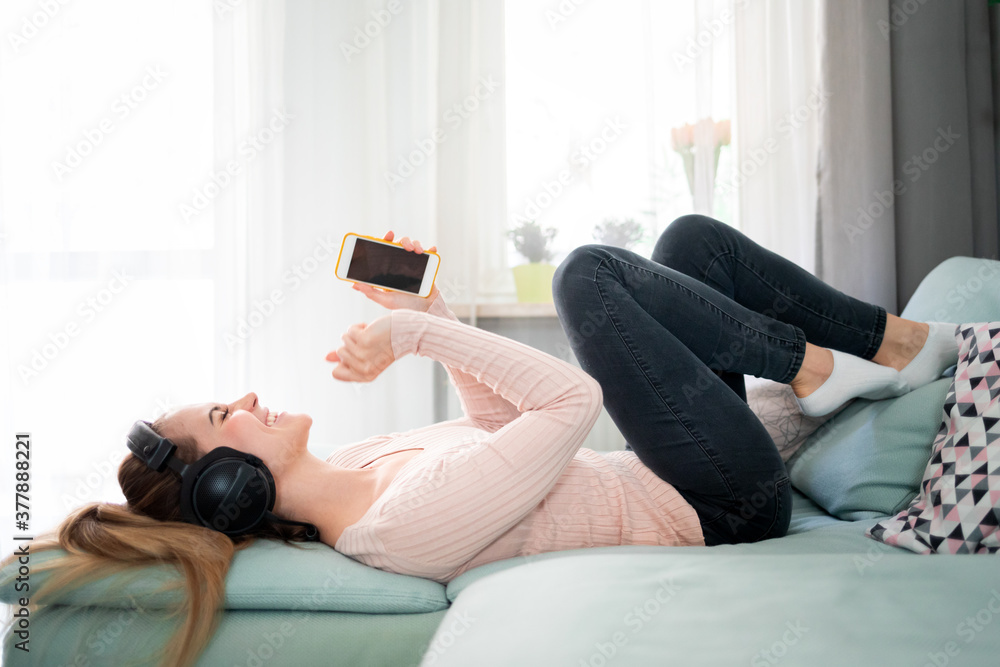 Relaxed woman with headphones and smartphone listening music while lying on comfortable sofa at home