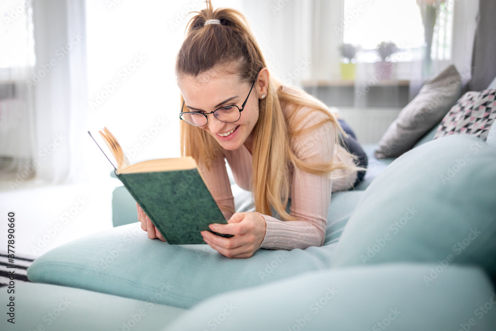 Happy woman with glasses reads book lying on comfortable sofa spending time at home