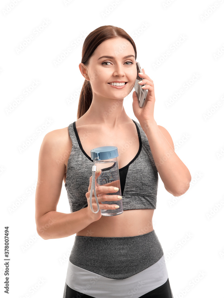 Sporty young woman with bottle of water talking by phone on white background