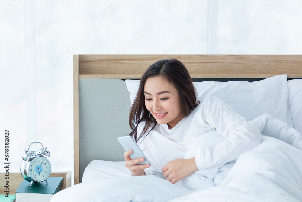 Happy young girl using a mobile phone lying on the bed at home in the morning. Girl playing with sma