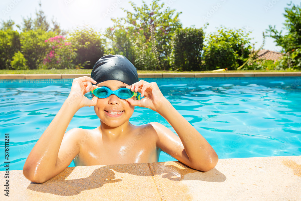 Happy little boy in swimming cap hold googles with hands on the border of outdoor pool look at camer