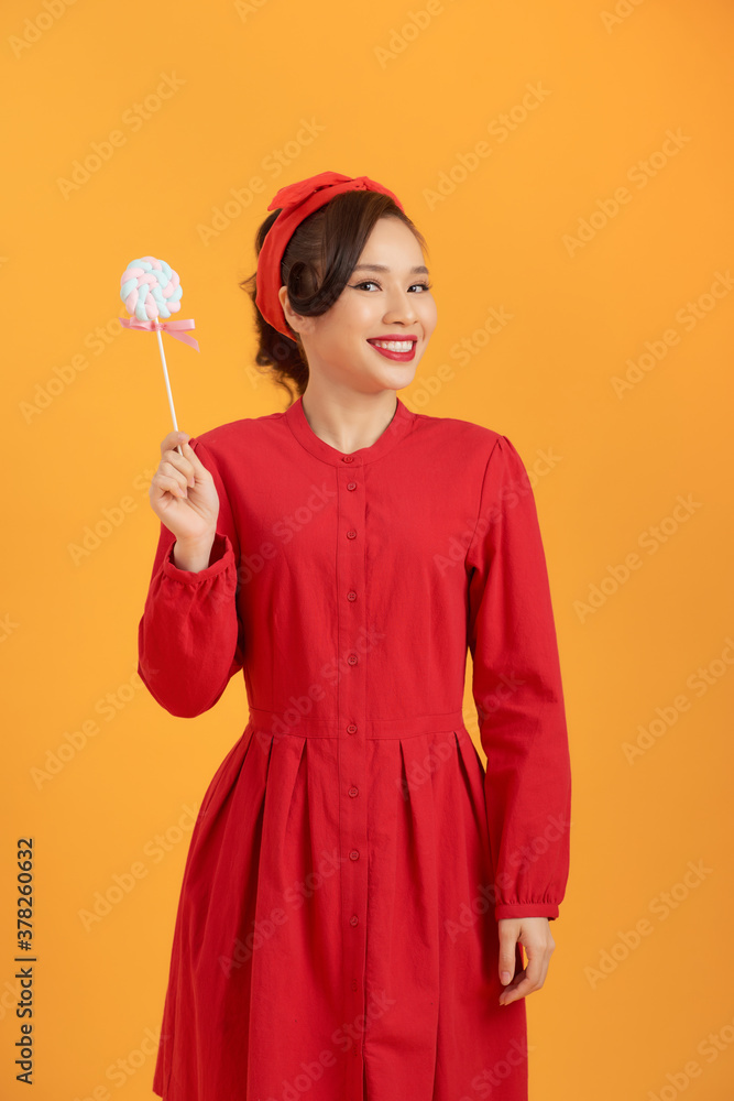 Smiling young Asian woman in red dress holding lollipop while standing over orange backgound.