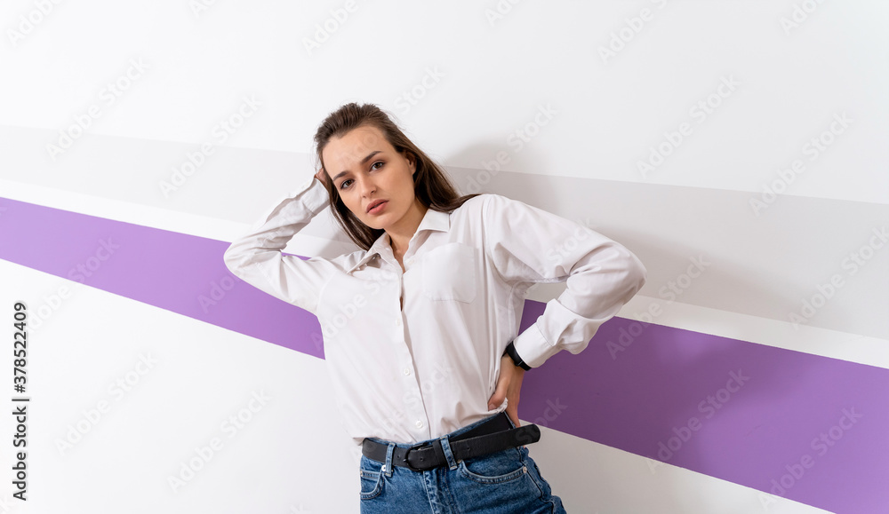 Full length photo of a girl in white shirt and blue jeans. Woman is standing near white wall with vi