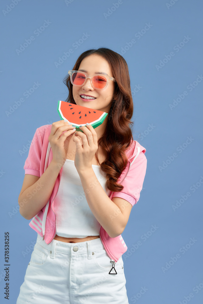 Cheerful young Asian woman holding watermelon and wearing sunglasses over blue background.