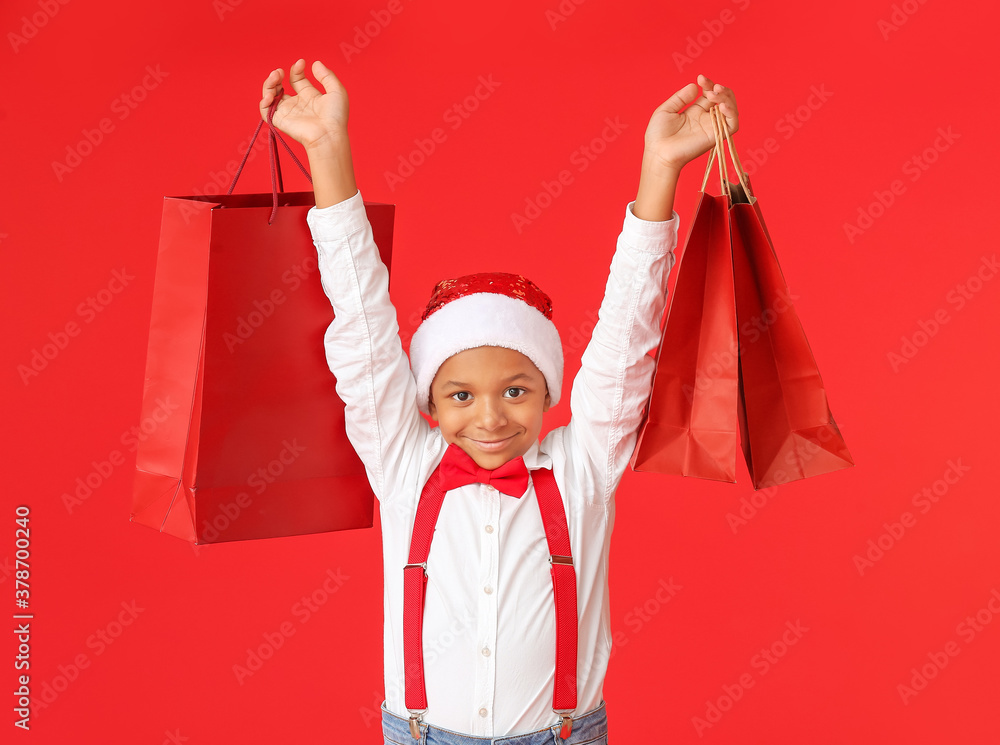 Cute African-American boy in Santa hat and with shopping bags on color background