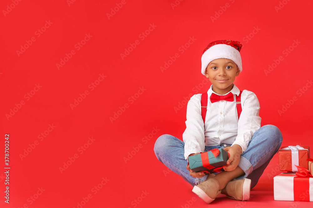 Cute African-American boy in Santa hat and with Christmas gifts on color background