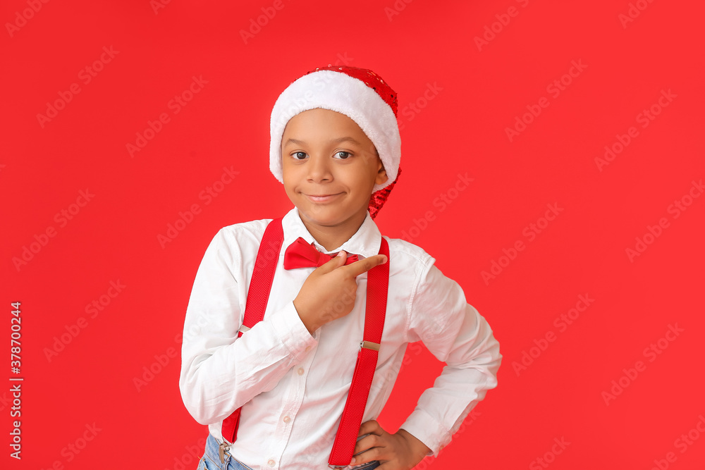 Cute African-American boy in Santa hat pointing at something on color background