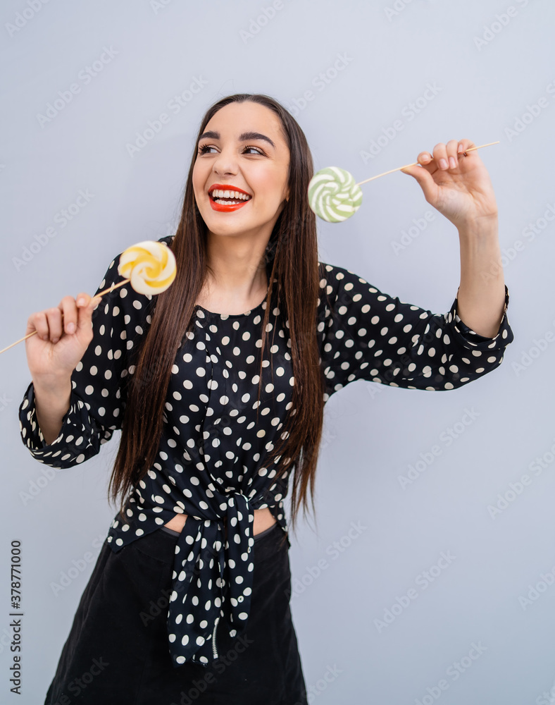 Young beauty in dotted shirt with two lollipops in hands. Cheerful lady with candies over grey backg