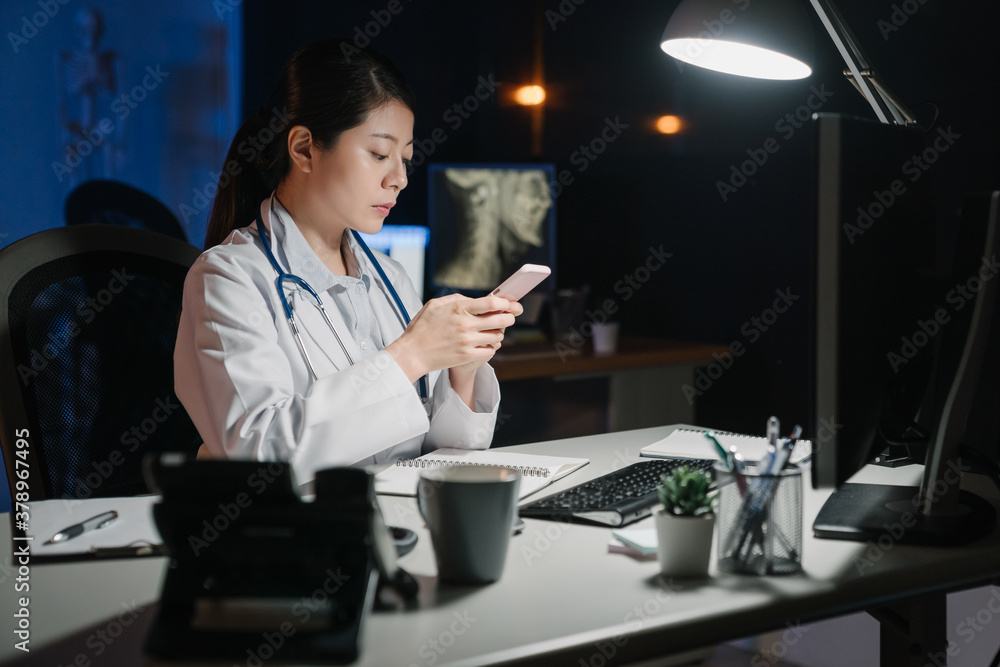 Shot of young asian chinese female doctor using mobile phone and text messaging while overtime worki