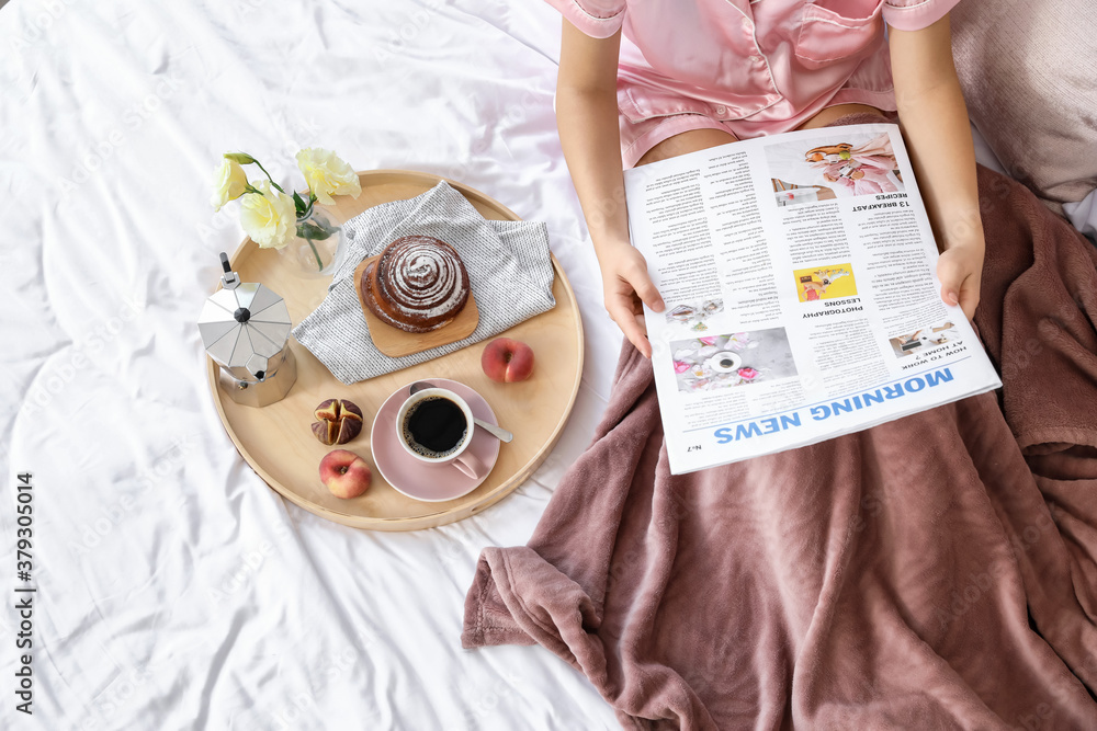 Young woman reading newspaper while having tasty breakfast in bed