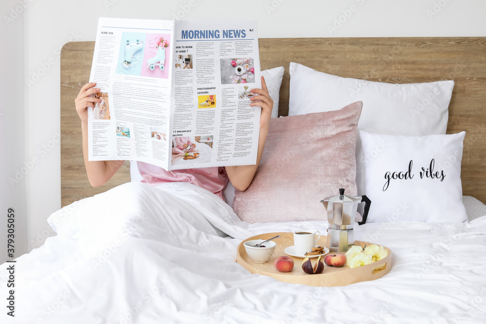 Young woman reading newspaper and having tasty breakfast in bed