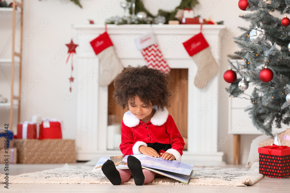 Cute African-American girl in Santa costume reading book at home on Christmas eve