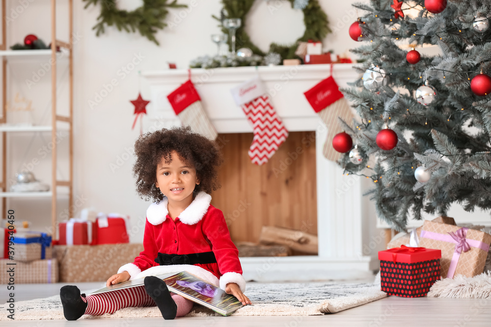 Cute African-American girl in Santa costume reading book at home on Christmas eve