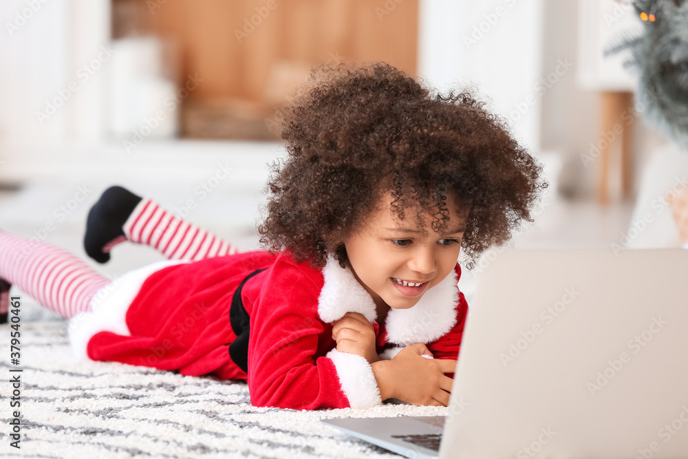 Cute African-American girl in Santa costume and with laptop at home on Christmas eve