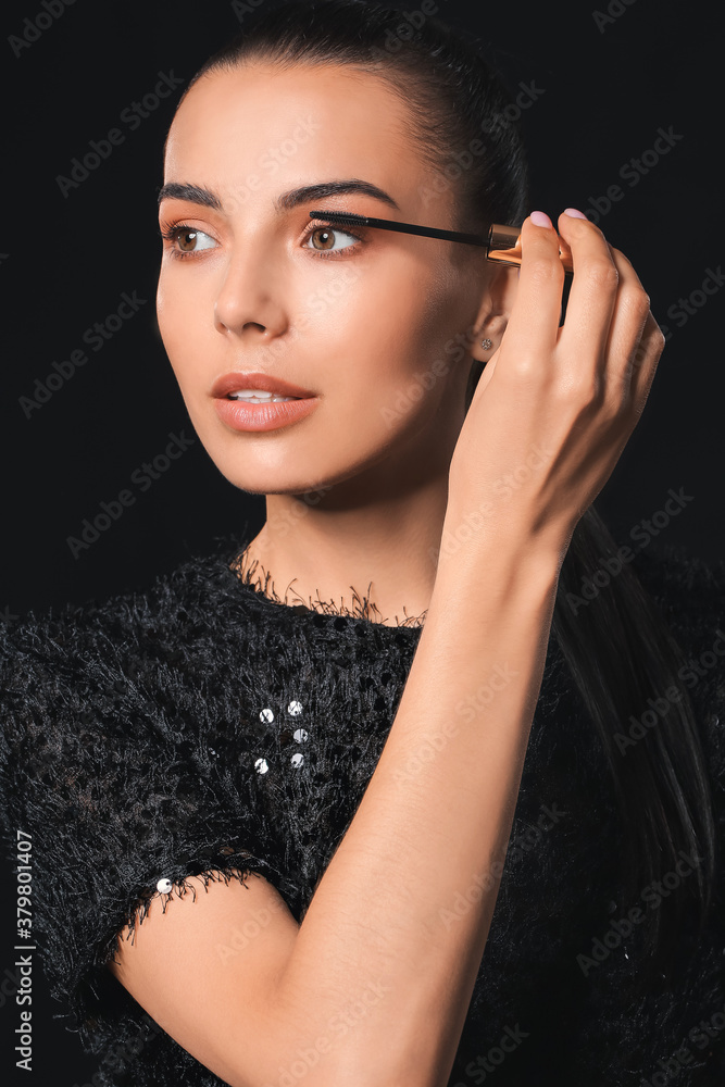 Beautiful young woman applying mascara against dark background