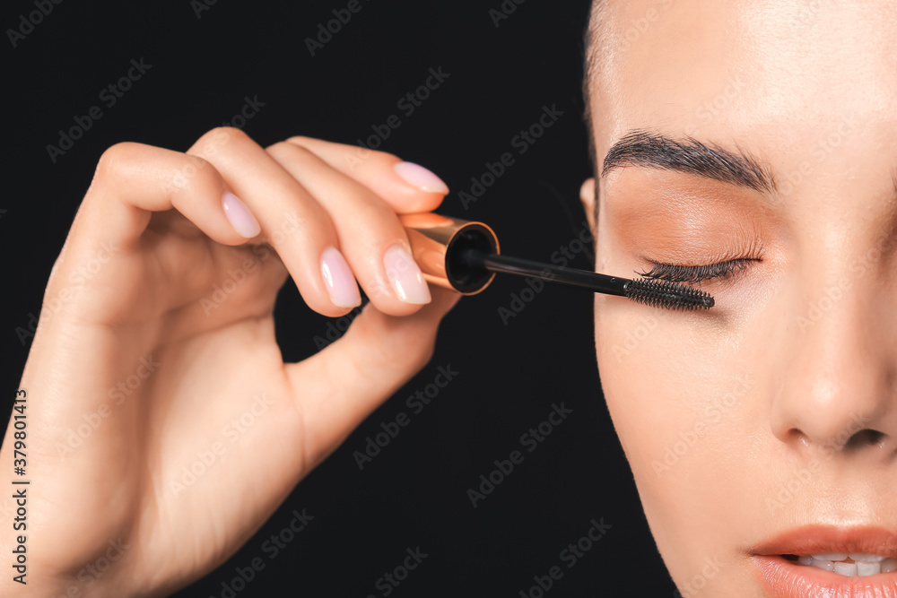 Beautiful young woman applying mascara against dark background, closeup