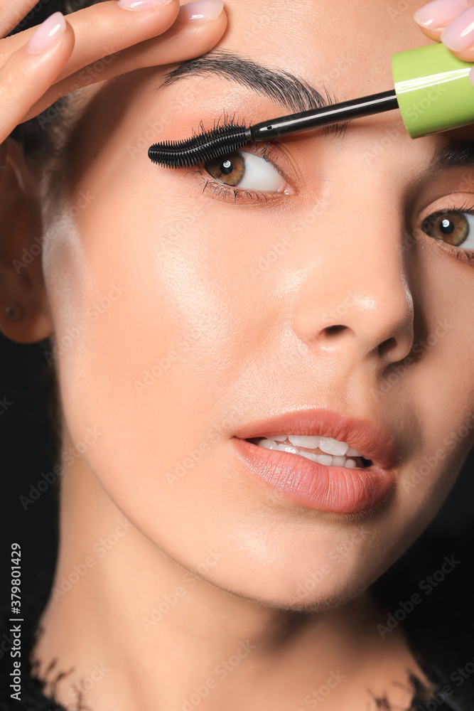 Beautiful young woman applying mascara against dark background, closeup