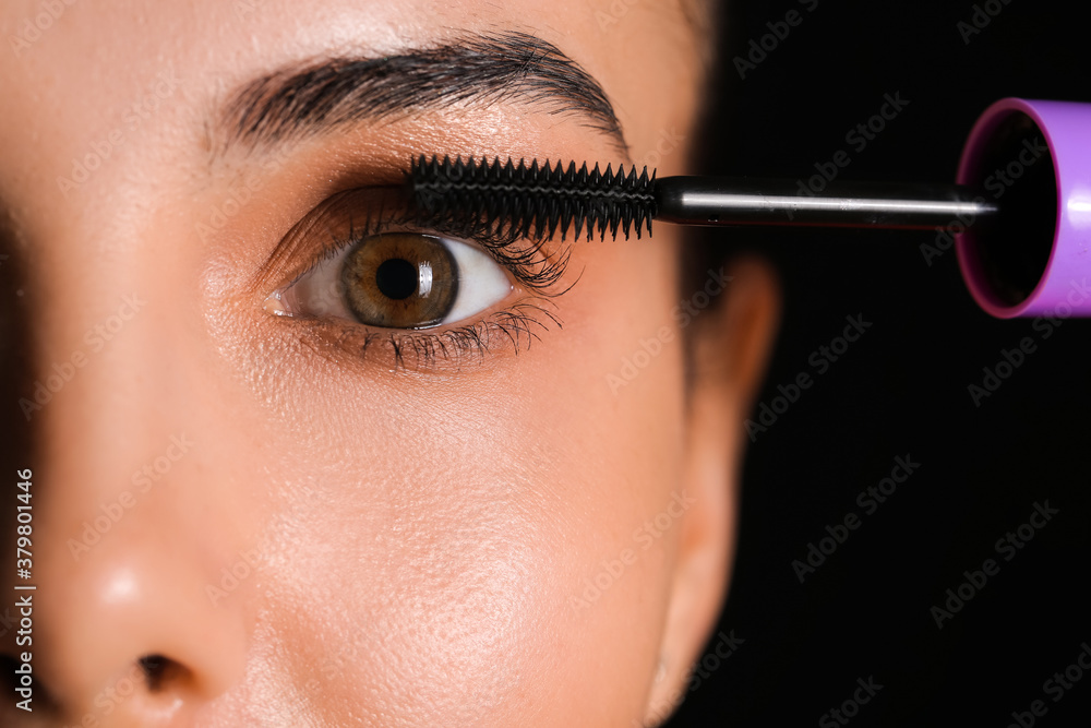 Beautiful young woman applying mascara against dark background, closeup