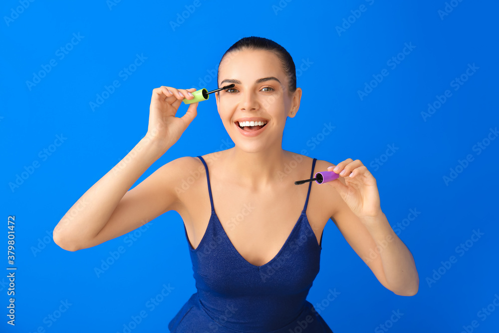 Beautiful young woman applying mascara on color background