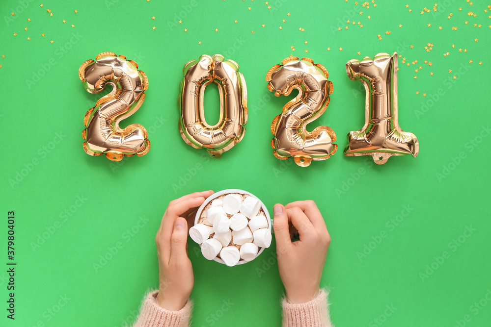 Hands with cup of hot chocolate and balloons in shape of figure 2021 on color background. New Year c