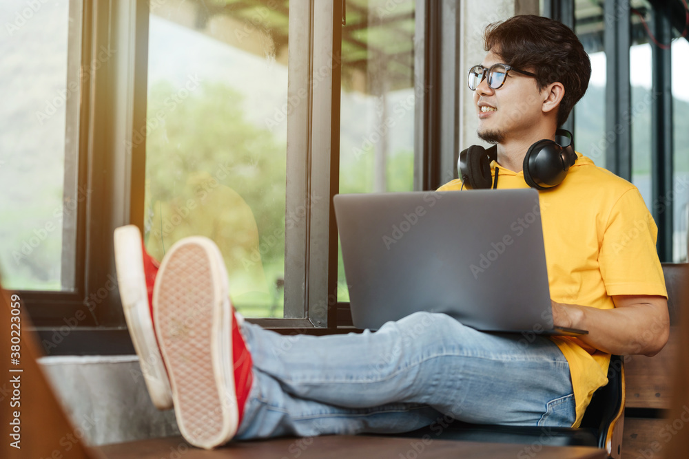 Happy Asian freelance man looking out of the window, thinking of content in social network via lapto