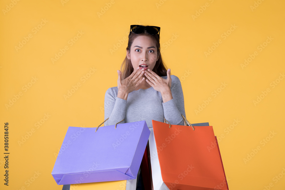 Beautiful young Asian woman carrying shopping bags looking surprised and happy isolated on yellow ba