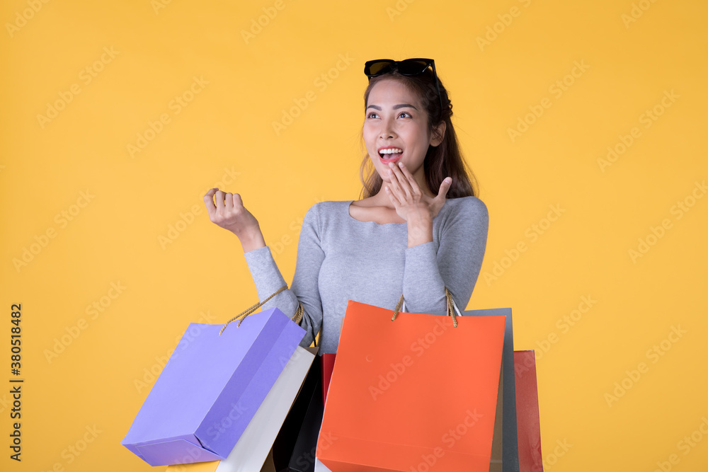 Beautiful young Asian woman carrying shopping bags looking surprised and happy isolated on yellow ba