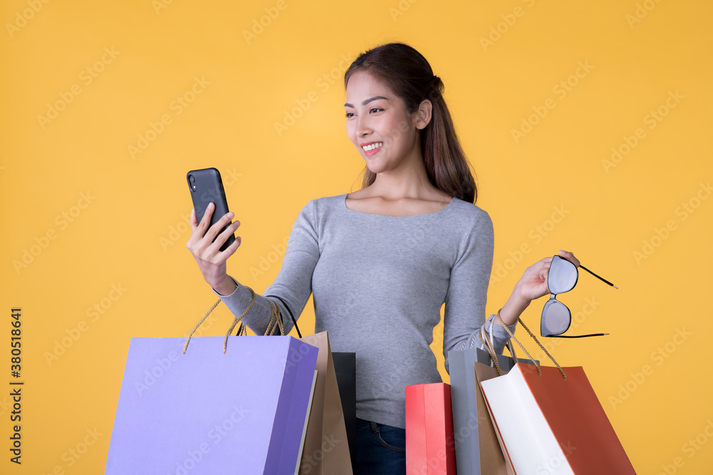 Beautiful young Asian woman with colourful shopping bags and smartphone isolated on yellow backgroun