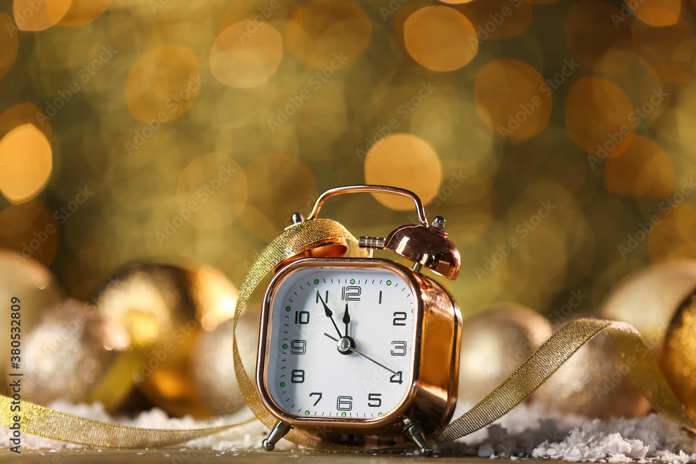 Alarm clock with ribbon and snow on table against blurred lights. Christmas time