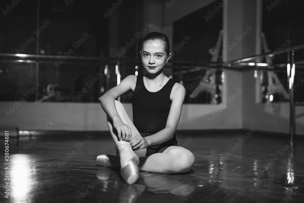 Portrait of pretty little ballerina sitting on floor and tying her pointe in classical dance studio.