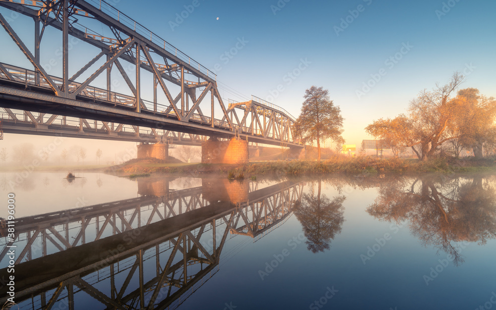 Beautiful railroad bridge and river in fog at sunrise in autumn. Industrial landscape with railway s