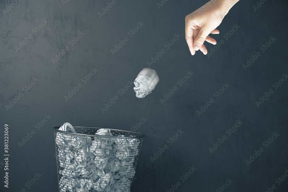 Hand trowing a paper into a wastebasket on gray concrete wall background.