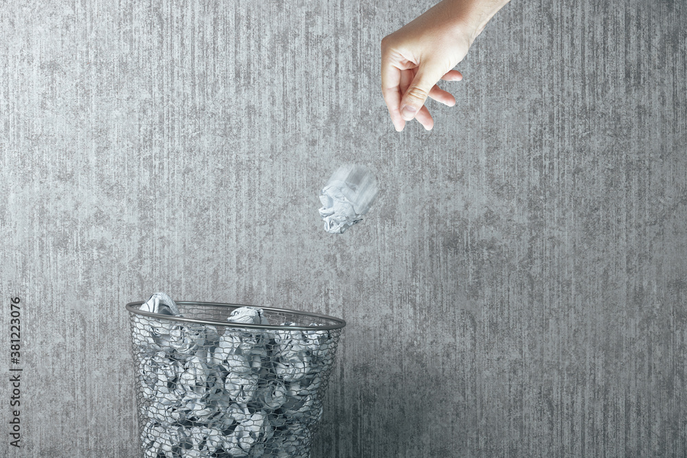 Hand trowing a paper into a wastebasket on concrete wall background.