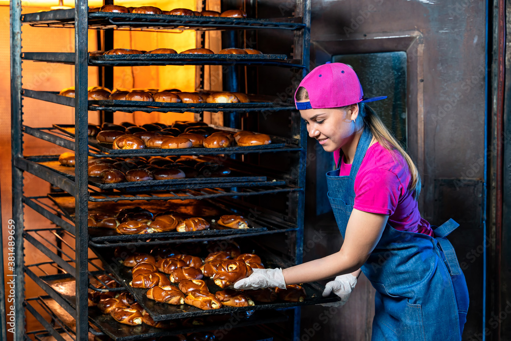 Young happy female worker in sterile clothes holding freshly baked muffins on tinplate inside food p